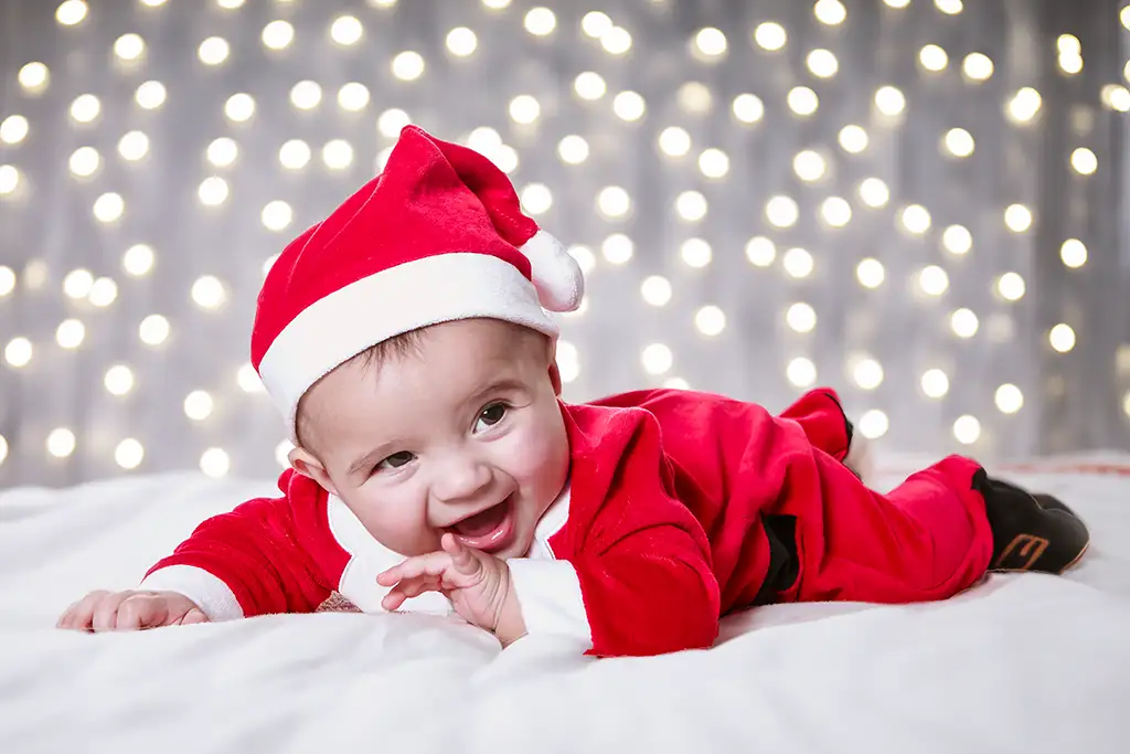 Fotografía de un bebé vestido como Papá Noel, con un fondo cálido de luces brillantes, perfecta para sesiones navideñas llenas de ternura.