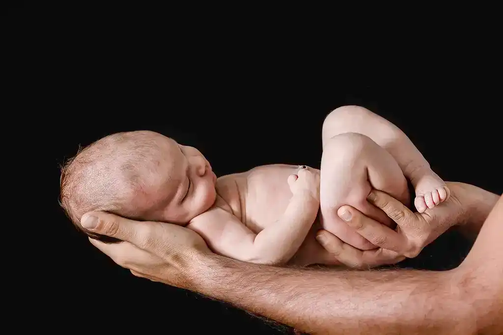 Fotografía artística de un recién nacido siendo sostenido delicadamente por las manos de su padre, destacando la conexión y protección paternal.