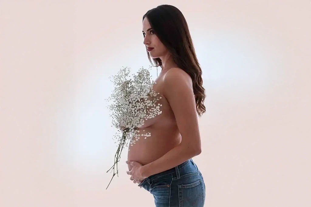Sesión de embarazo en estudio fotográfico de embarazo con fondo claro y un ramo de flores cubriendo el pecho.